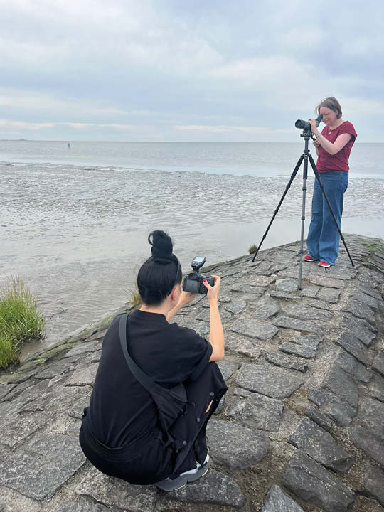 Silke schaut durch ihr Spektiv. Sie trägt ein bordeaux-rotes T-Shirt, eine Jeans und rote Sneaker. Vor ihr kniet die schwarz gekleidete Fotografin Alexandra Polina und fotografiert sie von unten hinauf. Beide sind auf einem Steinwall im Nordseewatt. 