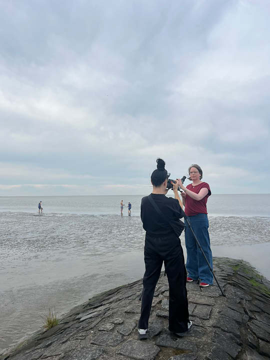 Silke steht aufrecht hinter ihrem Spektiv und schaut in die Ferne. Sie trägt ein bordeaux-rotes T-Shirt, eine Jeans und rote Sneaker. Vor ihr steht die schwarz gekleidete Fotografin Alexandra Polina und fotografiert sie. Beide sind auf einem Steinwall im Nordseewatt.